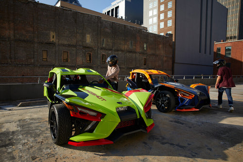 A bright green and bright blue and orange Polaris Slingshot on a rooftop in a city. Two men with helmets are getting into the Slingshots. 